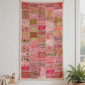 Könnte beinhalten: Ein rechteckiger Wandbehang mit einem Patchwork-Design aus verschiedenen rosa, roten und goldenen Stoffquadraten. Das Textil besteht aus mehreren bestickten und gemusterten Abschnitten, die mit sichtbaren weißen Nähten verbunden sind.