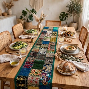 May include: A long rectangular table runner featuring a patchwork design of various embroidered fabric squares in shades of green, blue, gold, and black. The runner is centered on a wooden dining table set with plates, glasses, and napkins.