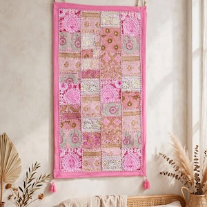 May include: A rectangular wall hanging featuring a patchwork design of various pink fabric squares. The piece is adorned with intricate embroidery, metallic thread accents, and small reflective embellishments. It has a solid pink border and two tassels at the bottom corners, suspended by a simple cord.