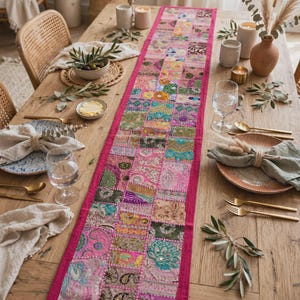 Peut inclure: Un long chemin de table rectangulaire composé de carrés de tissu patchwork dans des tons de rose, vert, doré et bleu. Le chemin de table est orné de broderies complexes, de paillettes et de petits miroirs, avec une bordure rose unie.