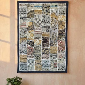 May include: A patchwork wall hanging with a dark blue border. The design is made up of numerous fabric squares in beige, brown, and navy blue, each with a different pattern. The wall hanging is displayed on a light pink wall.