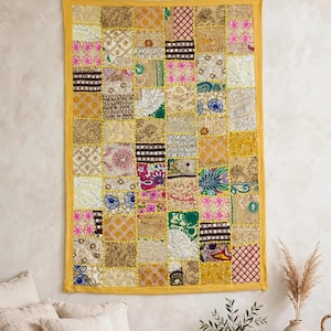 Op de afbeelding: Een rechthoekige wandhanger met een patchworkontwerp van diverse stoffen vierkanten in goud-, geel-, roze-, groen- en wittinten. Het textiel vertoont ingewikkeld borduurwerk, lovertjes en patronen van metaaldraad met een gele rand.