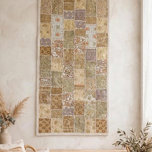 May include: A rectangular wall hanging featuring a patchwork design of various fabric squares in shades of gold, olive green, and muted purple. The textile is decorated with intricate embroidery, metallic thread accents, and small sequins.