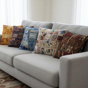 Op de afbeelding: Een lichtgrijze bank met vijf decoratieve kussens. De kussens hebben verschillende kleurrijke patchwork-ontwerpen in geel, paars, blauw en bruin. De kamer heeft een neutraal kleurenpalet met een houten salontafel.