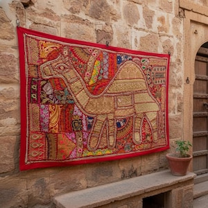 May include: A vibrant, red-bordered tapestry featuring a camel design, hangs on a stone wall. The camel is rendered in gold and tan hues, with intricate patterns. The scene includes a wooden door and a small potted plant, suggesting an outdoor setting.