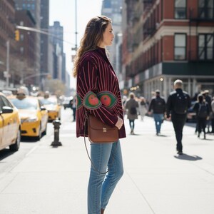 Pode incluir: Uma pessoa vestindo uma jaqueta bord&ocirc; com listras rosa verticais e desenhos verdes e laranja em forma de olho. Ela est&aacute; usando jeans azul claro, botas marrons e carregando uma bolsa transversal de couro marrom. O fundo mostra uma rua da cidade com t&aacute;xis amarelos.