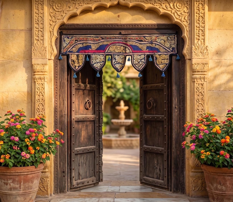 May include: An open, weathered wooden door with ornate carvings and a decorative textile valance. The doorway leads to a courtyard with a fountain. Potted plants with pink, orange, and yellow flowers flank the entrance.