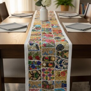 May include: A vibrant patchwork table runner with a white border, placed on a dark wooden table. The runner showcases embroidered squares in blue, pink, yellow, and green. A white vase with flowers is in the centre of the table.