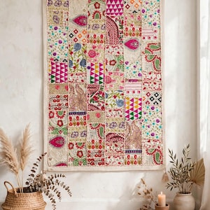 Op de afbeelding: Een rechthoekige wandhanger met een patchworkontwerp van diverse geborduurde stoffen vierkanten. Het textiel toont een levendig kleurenpalet met roze, groen, rood en goud, voorzien van ingewikkelde bloemen-, geometrische en paisley-patronen.