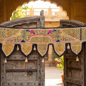 May include: A decorative textile door hanging with a gold and black design. The textile features floral and geometric patterns with pink, blue, and green accents. The hanging has a scalloped bottom edge with tassels. The door is made of weathered wood.