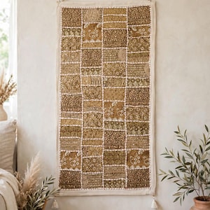 Puede incluir: Un tapiz rectangular con un diseño de parches de varios cuadrados de tela en tonos oro y verde oliva. La pieza está adornada con bordados intrincados, lentejuelas e hilos metálicos, rematada con un borde blanco y dos pequeñas borlas colgantes.