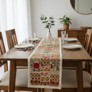 Könnte beinhalten: Ein Holztisch mit einem dekorativen Tischläufer mit Blumenmuster. Der Läufer hat ein Patchwork-Design mit roten, rosa und grünen Blumenmustern. Der Tisch ist mit Tellern, Gläsern und Besteck gedeckt.