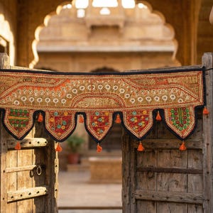 May include: An ornate, embroidered textile hangs above a weathered wooden door. The textile features intricate patterns in shades of orange, red, and green, with decorative tassels. The scene is framed by stone architecture and flowering plants.