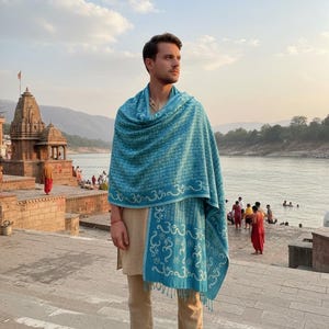 Può includere: Una persona indossa uno scialle turchese con un motivo ripetuto e frange. Lo scialle è drappeggiato su una tunica e pantaloni beige. Lo sfondo include un fiume, un tempio e persone.