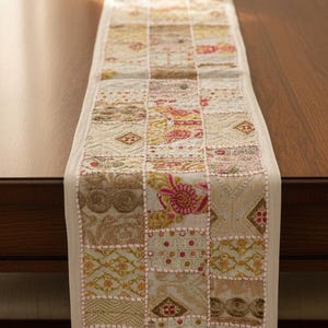 May include: A rectangular table runner with a patchwork design. The runner features squares of various patterns in beige, pink, and gold tones. The runner is placed on a wooden table. A white vase with white flowers sits on the table.