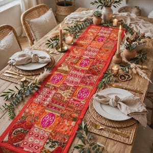 May include: A long rectangular table runner featuring a vibrant patchwork design of red, orange, and pink fabric squares. The runner is decorated with intricate embroidery, metallic thread accents, and small circular mirrors, creating a textured surface.
