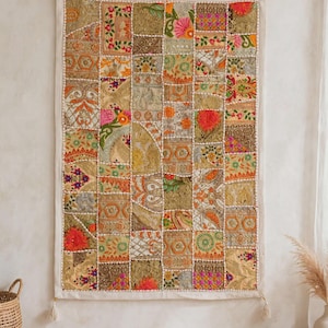 May include: A rectangular wall hanging featuring a patchwork design of various fabric squares in shades of gold, orange, pink, and green. The textile displays intricate embroidery, metallic threads, and small decorative tassels at the bottom corners.