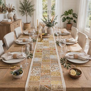 May include: A rectangular table runner featuring a patchwork design of embroidered fabric squares in shades of gold, yellow, and cream. The runner is centered on a long wooden dining table set with white plates, linen napkins, and gold cutlery.