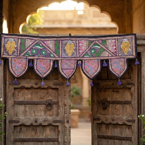 May include: A colorful, embroidered textile door hanging with a geometric design, featuring pink, green, and yellow accents. The hanging is adorned with purple tassels and hangs over a weathered wooden door. Potted plants frame the doorway.