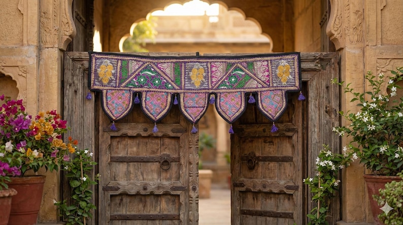 May include: A colorful, embroidered textile door hanging with a geometric design, featuring pink, green, and yellow accents. The hanging is adorned with purple tassels and hangs over a weathered wooden door. Potted plants frame the doorway.
