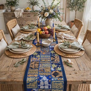 May include: A rectangular wooden dining table set with a blue patchwork table runner featuring intricate gold and floral embroidery. The table includes woven placemats, ceramic plates, linen napkins with olive sprigs, and a central bowl of fruit.