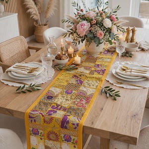 May include: A rectangular table runner featuring a vibrant patchwork design of yellow, gold, and purple fabric squares. Each square displays intricate embroidered floral, paisley, and geometric patterns with metallic thread accents and a solid yellow border.