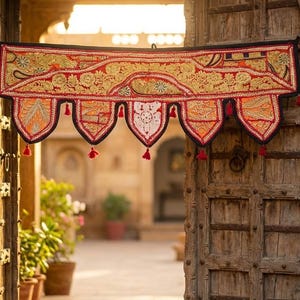 May include: A decorative textile valance with intricate embroidery in red, gold, and orange hues hangs above a weathered wooden door. The valance features a scalloped edge with red tassels. The scene is framed by a stone archway and potted plants.