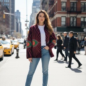 Pode incluir: Cardig&atilde; bord&ocirc; com um design de olho em tons de verde, vermelho e rosa. Usado sobre uma camiseta branca, combinado com jeans azuis e botas marrons. O fundo mostra uma rua da cidade com t&aacute;xis amarelos e pedestres.