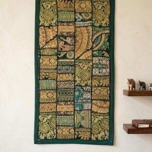 Puede incluir: Un tapiz rectangular con un diseño de patchwork. El tapiz presenta un borde verde oscuro y un panel central compuesto por varios cuadrados en tonos dorados, verdes y turquesas. Intrincados patrones bordados adornan cada cuadrado, creando una apariencia rica y texturizada.