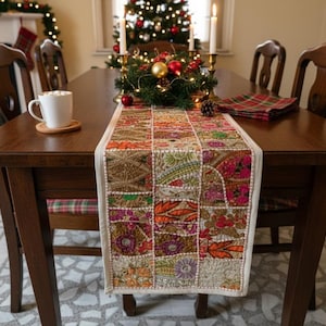 May include: A wooden dining table set for a holiday meal. A colorful patchwork table runner is centered on the table. A white mug sits on a coaster. A Christmas tree and garland decorate the background.