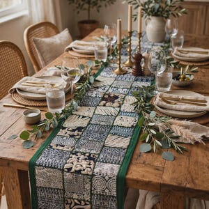 May include: A rectangular table runner featuring a patchwork design of various blue, white, and silver embroidered fabric squares with a solid green border, laid across a wooden dining table set with plates, glasses, candles, and greenery.