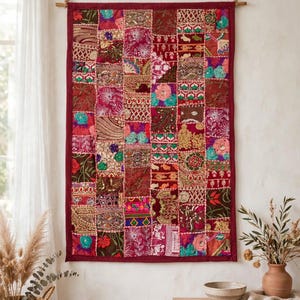 Peut inclure: Une tenture murale rectangulaire composée d'un patchwork de carrés de tissu dans des tons rouge profond, magenta et or. Le textile présente des broderies complexes, des paillettes et des motifs floraux, suspendu à une tringle en bois avec une corde.