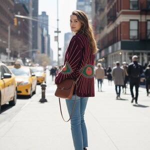 Pode incluir: Uma pessoa est&aacute; numa rua da cidade usando um su&eacute;ter bord&ocirc; com listras rosa e desenhos em forma de olho. Ela usa jeans azuis, botas marrons e carrega uma bolsa de couro marrom. T&aacute;xis amarelos e edif&iacute;cios ao fundo.