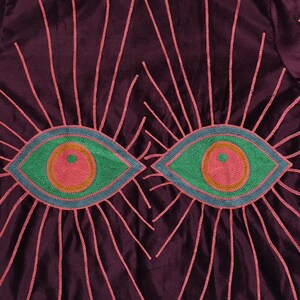 Pode incluir: Close-up de um tecido roxo escuro com desenhos de olhos bordados. Cada olho &eacute; verde com um centro rosa e contorno azul, rodeado por linhas rosa radiantes. O tecido tem um brilho subtil.