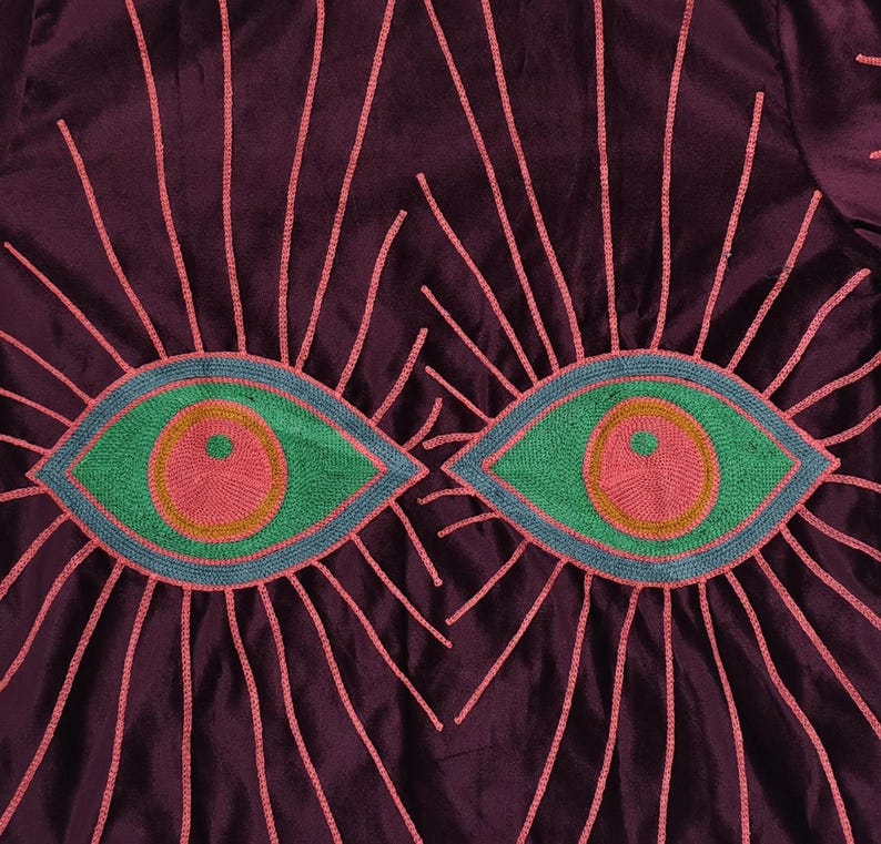 Pode incluir: Close-up de um tecido roxo escuro com desenhos de olhos bordados. Cada olho &eacute; verde com um centro rosa e contorno azul, rodeado por linhas rosa radiantes. O tecido tem um brilho subtil.