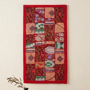 Puede incluir: Un tapiz de pared rectangular rojo con un diseño de patchwork. El patchwork presenta varios patrones en rojo, verde, blanco y dorado. El tapiz está enmarcado con un borde rojo liso. Un pequeño jarrón negro con una ramita de vegetación y una pila de libros están en primer plano.