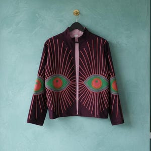 Pode incluir: Casaco borgonha com z&iacute;per e design de olho &uacute;nico. O casaco apresenta um z&iacute;per central, gr&aacute;ficos de olhos rosa e verdes na frente e nas mangas e linhas rosa radiantes. Pendurado em um cabide preto contra uma parede verde-azulado.