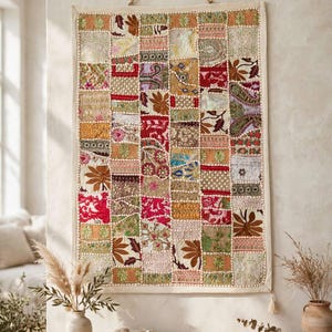 Puede incluir: Un tapiz rectangular con un diseño de retazos de varios cuadrados de tela bordados en tonos rojo, dorado, verde y crema. El textil está suspendido por un cordón fino y muestra intrincados patrones florales y geométricos.