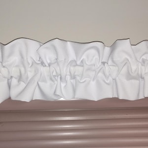 May include: A white ruffled valance, likely for a window treatment, is displayed. The valance is made of a soft, white fabric with a gathered design. The valance is positioned above a set of horizontal blinds, creating a layered window decor.