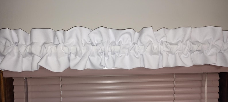 May include: A white ruffled valance, likely for a window treatment, is displayed. The valance is made of a soft, white fabric with a gathered design. The valance is positioned above a set of horizontal blinds, creating a layered window decor.