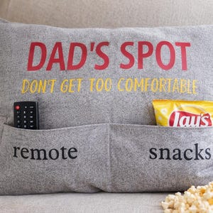 May include: Gray pillow with pockets labeled "remote" and "snacks." The pillow has the text "DAD'S SPOT" in red and "DON'T GET TOO COMFORTABLE" in yellow. A remote and a bag of chips are in the pockets.