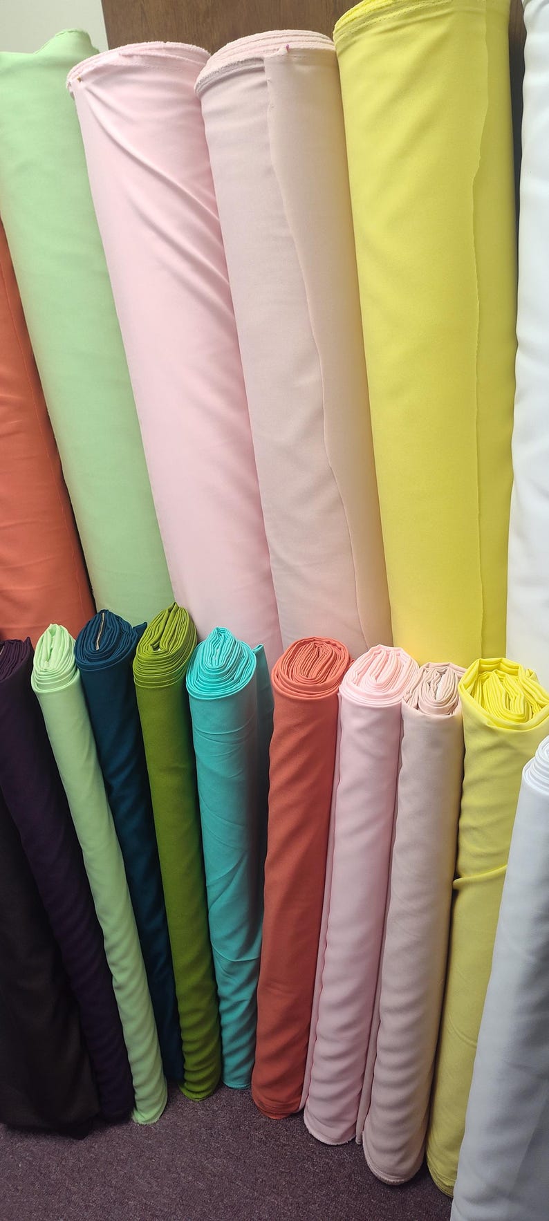 May include: A collection of fabric rolls in various colors, including green, pink, yellow, and teal. The rolls are tightly wound and stacked together, showcasing a variety of textiles for sewing and crafting projects.