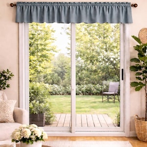 May include: A blue valance hangs above a sliding glass door, framed by a white trim. The door opens to a backyard with a wooden deck, a chair, and lush greenery. A round wooden coffee table and a beige rug are in the foreground.
