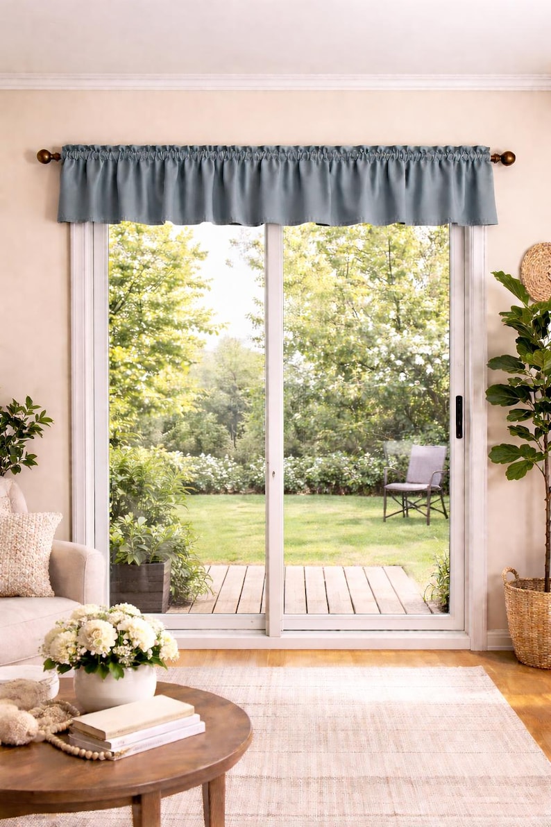 May include: A blue valance hangs above a sliding glass door, framed by a white trim. The door opens to a backyard with a wooden deck, a chair, and lush greenery. A round wooden coffee table and a beige rug are in the foreground.