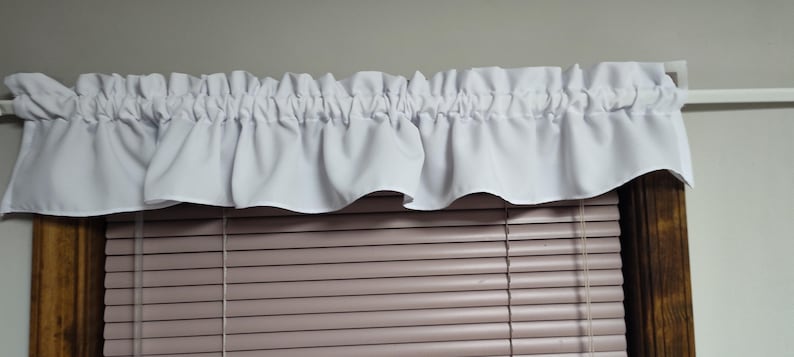 May include: White ruffled valance over a window with closed blinds. The valance is made of a soft, flowing fabric and is gathered at the top. The blinds are a light brown color.