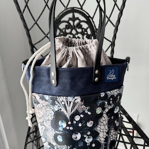 Peut inclure: Un sac fourre-tout artisanal avec un dessus en daim bleu marine et une base à imprimé floral. Le sac est doté de poignées en cuir noir et d'une fermeture à cordon. Le sac porte une petite étiquette indiquant "Crazy Designs".