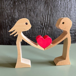 May include: Two wooden figures holding a red heart between them. The figures are stylized and have simple features. The heart is a separate piece and is positioned between their outstretched arms.
