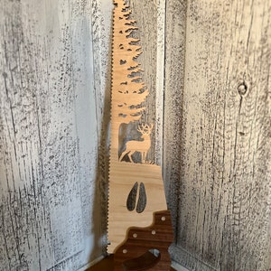 May include: A wooden saw blade with a silhouette of a pine tree, a deer, and a deer track. The handle is made of a darker wood.