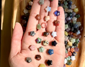 Op de afbeelding: Een verscheidenheid aan stervormige edelstenen in verschillende kleuren en texturen, gepresenteerd in een houten kom en in de open hand van een persoon. De edelstenen omvatten tinten blauw, groen, roze en goud.