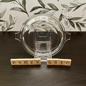 May include: A clear glass Pyrex baking dish lid with a handle on each side. The lid is sitting on a wooden surface with the letters PYREX and 681C spelled out in wooden tiles.